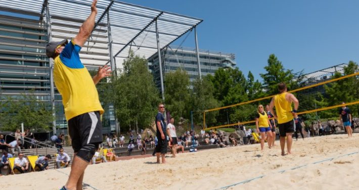A team of players is playing beach volleyball on the beach under a sunny sky, using a yellow volleyball and net.