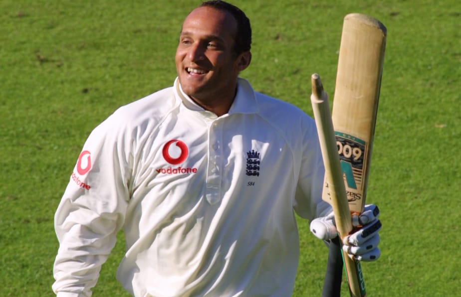 A person in a sports uniform is holding cricket equipment while standing on a grassy outdoor field.