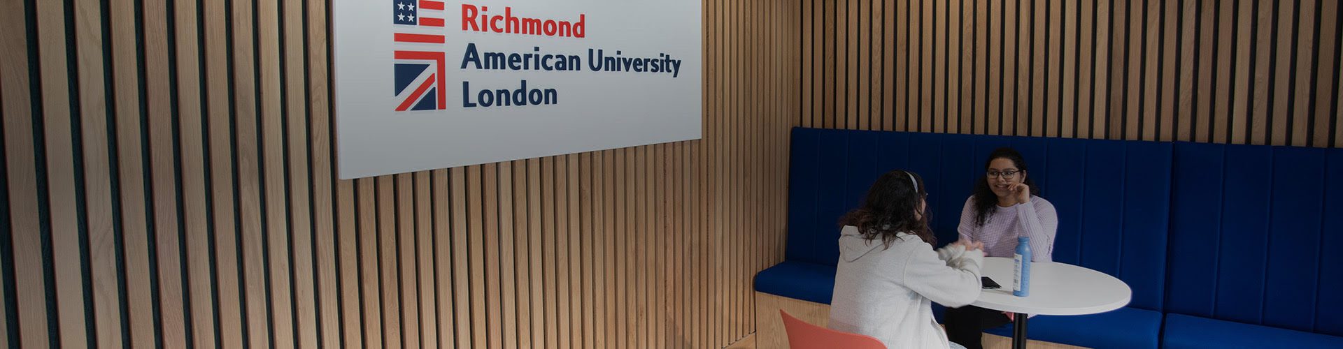 Two people are seated opposite each other at a white table inside a modern room with wooden slats and a sign for 快活视频 American University London.
