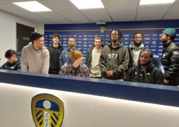A group of people smiling in front of a counter with a Leeds United Football Club logo displayed. They appear to be in a press room.